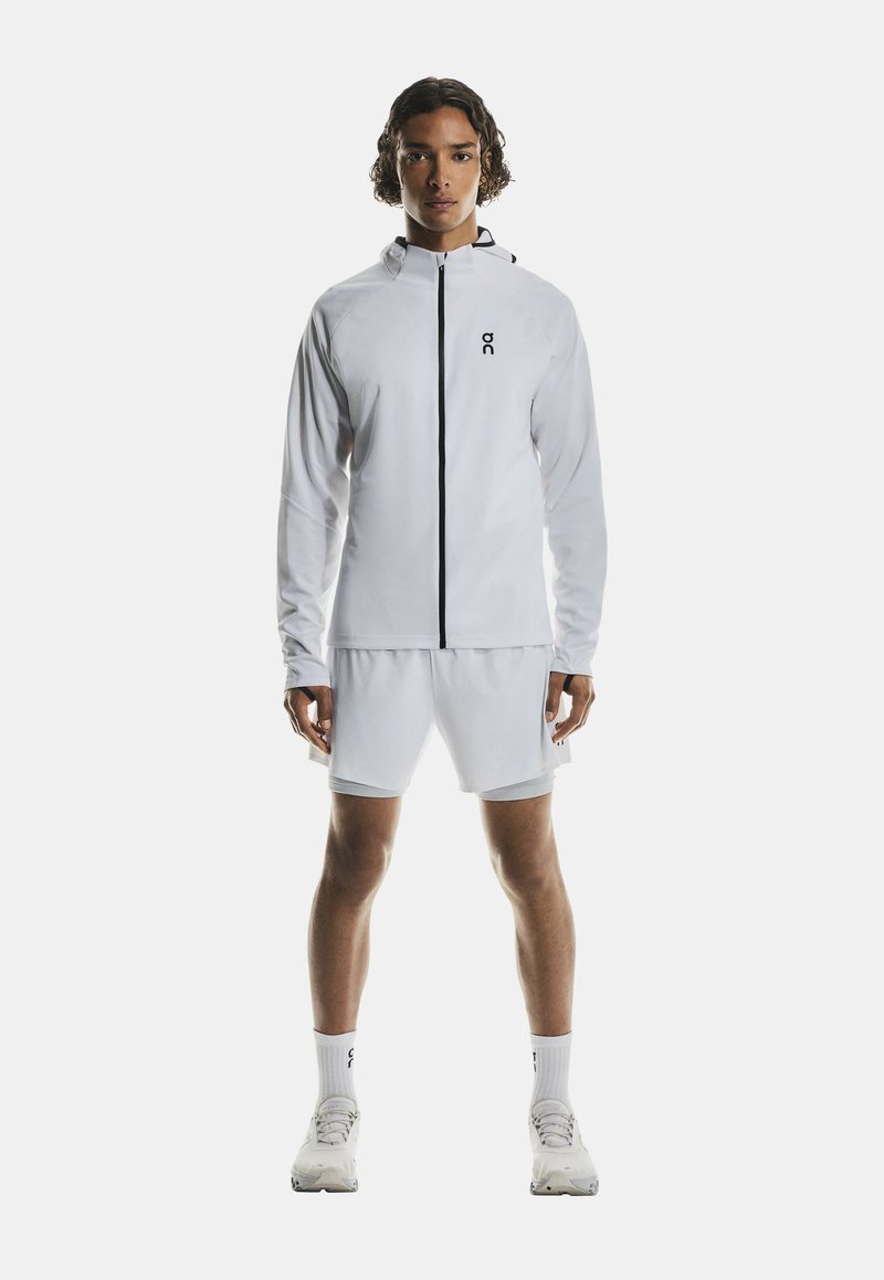 Veste de sport blanche avec une fermeture éclair noire, assortie à un short blanc. Tissu lisse, design minimaliste, avec un logo sur la poitrine.