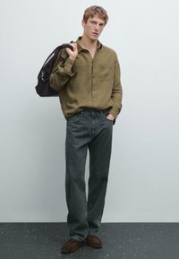 Giovane uomo che indossa una camicia verde oliva e jeans neri larghi, con uno zaino nero a tracolla, in piedi davanti a un muro grigio semplice.
