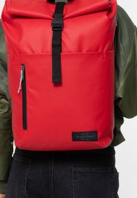 Zaino rosso a serratura superiore realizzato in materiale resistente, con una tasca frontale con zip, accessori neri e una patch con logo sulla parte anteriore inferiore.