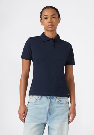 Donna con capelli intrecciati che indossa una polo blu navy a maniche corte e jeans azzurri a vita alta, in piedi davanti a uno sfondo semplice.