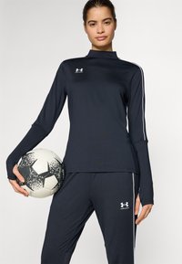 Dark blue athletic top with long sleeves, high collar, white side stripes, paired with matching pants. Holds a black and white soccer ball.