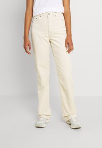 Cremfarbene Cordhosen mit hohem Bund und geradem Schnitt. Verfügt über Taschen und einen Knopfverschluss. Kombiniert mit hellen Turnschuhen.