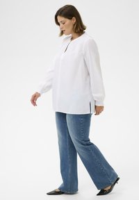 Blouse blanche à manches longues avec un col rond et des fentes latérales, associée à un jean évasé bleu et des escarpins noirs à bout pointu. Texture de tissu lisse.