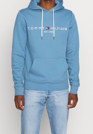 Osoba nosząca niebieską bluzę z kapturem Tommy Hilfiger z białymi sznureczkami i jasnoniebieskie dżinsy, widok z przodu pokazujący logo i przednią kieszeń.