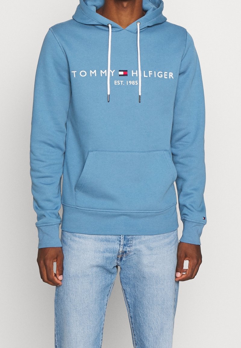 Personne portant un sweat à capuche bleu Tommy Hilfiger avec des cordons blancs et un jean bleu clair, vue de face montrant le logo et la poche avant.