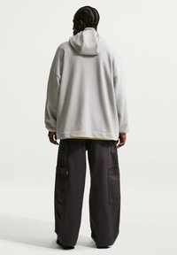 Personne aux cheveux tressés portant un sweat à capuche gris clair surdimensionné et un pantalon cargo noir ample, debout de dos sur un fond uni.