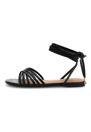 Schwarze flache Sandale mit mehreren dünnen Frontriemen und einer Schnürung am Knöchel, die mit einem kleinen dekorativen Anhänger an der Ferse verziert ist.