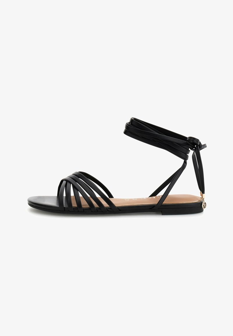 Sandale plate noire avec plusieurs fines brides avant et fermeture à nouer autour de la cheville, ornée d'un petit charm décoratif au talon.