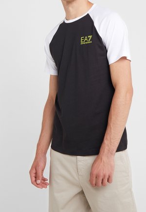 Homme portant un t-shirt à manches courtes noir et blanc avec un logo vert EA7 Emporio Armani, et un pantalon beige, présenté du cou aux hanches.