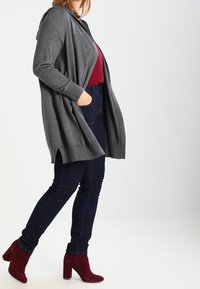 Szary, dzianinowy kardigan z kapturem, otwarty z przodu i z bocznymi rozcięciami, w połączeniu z ciemnoniebieskimi dżinsami i burgundowymi botkami na słupku.