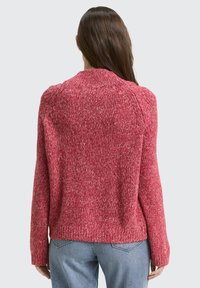 Frau mit langen braunen Haaren, die einen roten Strickpullover und hellblaue Jeans trägt, mit dem Rücken zur Kamera vor einem schlichten Hintergrund.