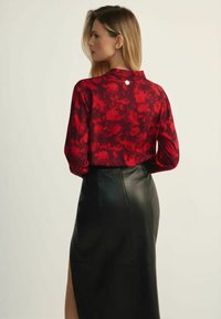 Kobieta z długimi blond włosami ma na sobie czerwoną bluzkę w wzory oraz czarną skórzaną spódnicę z tylnym rozcięciem, stoi bokiem na jednolitym tle.