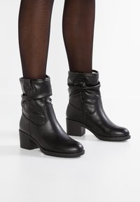 Bottes cheville en cuir noir avec tige souple, fermeture éclair latérale et talon bloc de 3 pouces, présentant une texture lisse et brillante.