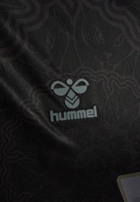 Czarna tkanina z subtelnymi geometrycznymi wzorami oraz szarym logo marki hummel z stylizowanym kształtem pszczoły na środku.