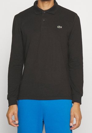 Mężczyzna w czarnej koszulce polo z długim rękawem, z małym zielonym logo krokodyla na piersi oraz w jaskrawo niebieskich spodniach.