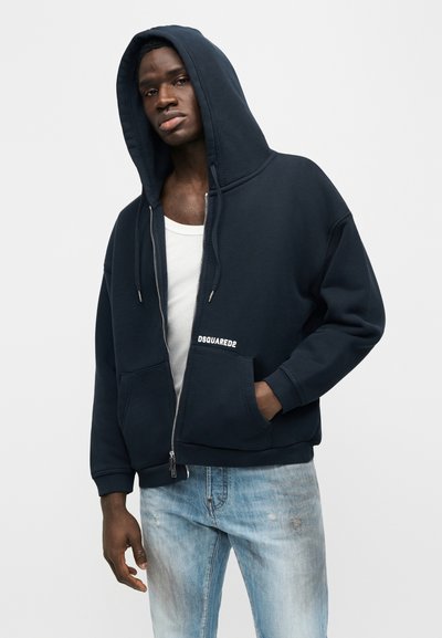 Άνδρας που φοράει μπλε σκούρο φούτερ με φερμουάρ και κουκούλα σηκωμένη, λευκό αμάνικο μπλουζάκι και ανοιχτόχρωμο ξεβαμμένο τζιν, στέκεται μπροστά σε απλό φόντο.