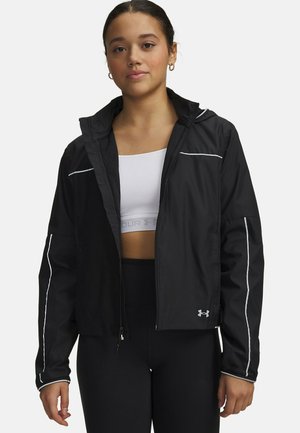 Young woman wearing a black zip-up jacket with reflective stripes, white sports bra, and black leggings against a plain white background.