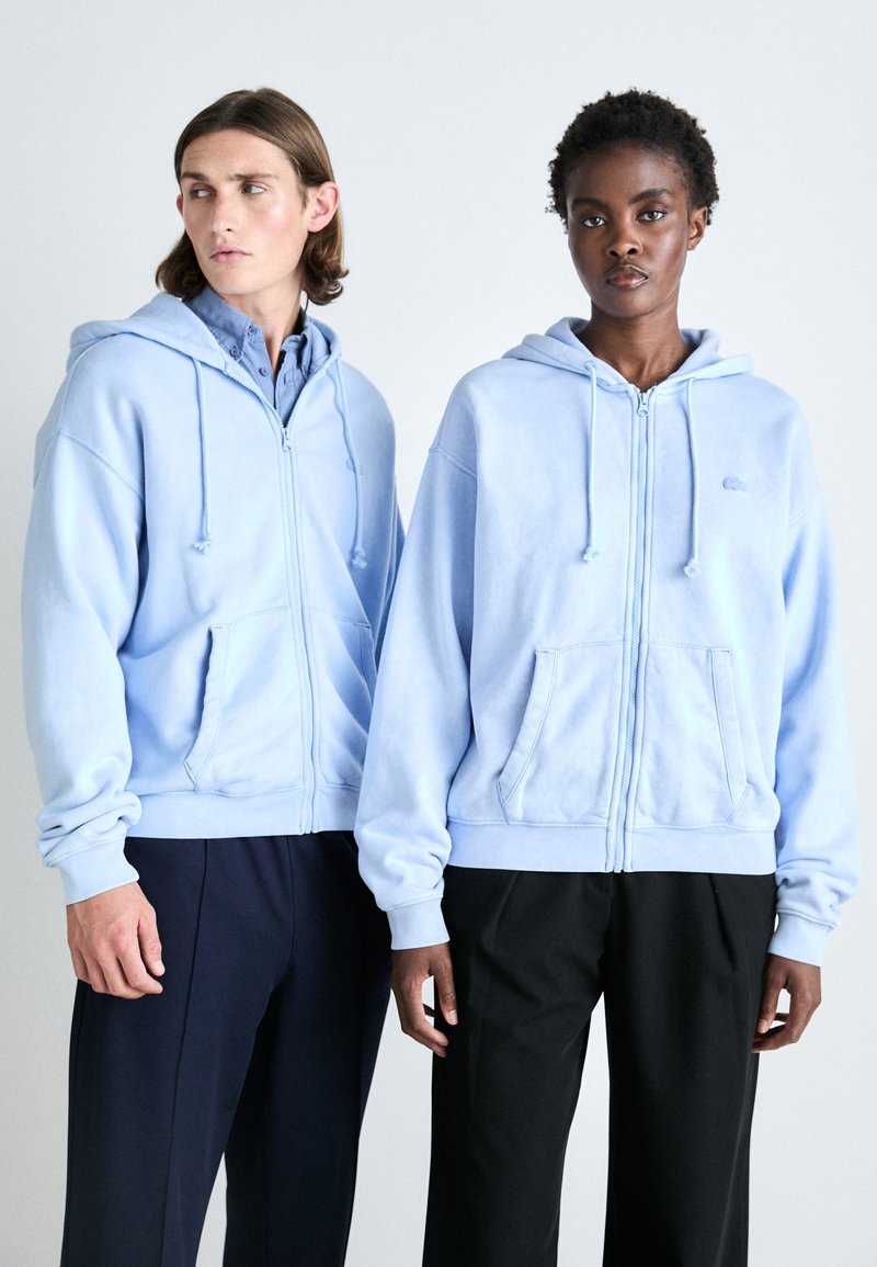 Sudaderas con cremallera de color azul claro, fabricadas en tejido suave, que presentan bolsillos tipo canguro, cordones ajustables y puños acanalados. Corte ajustado y mínima marca.