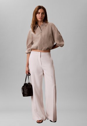 Femme portant une veste courte beige zippée et un pantalon blanc taille haute à jambes larges, tenant un sac seau en cuir noir, debout sur fond gris.