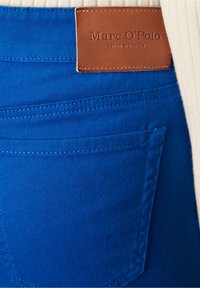 Jeans de mezclilla azul con bolsillos traseros, que presentan una etiqueta de marca de cuero marrón. La tela tiene una textura suave con un acabado ligeramente brillante.