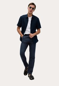 Marineblaues Kurzarmhemd über einem weißen T-Shirt, kombiniert mit dunklen Hosen und schwarzen Sandalen. Das Hemd hat ein lässiges Design und eine Knopfleiste.