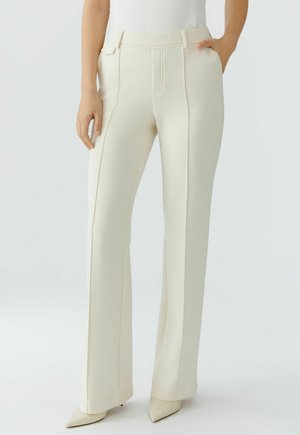 Frau trägt hoch taillierte, cremefarbene, maßgeschneiderte Hosen mit vorderen Nähten und spitzen Absätzen, steht mit einer Hand in der Tasche.