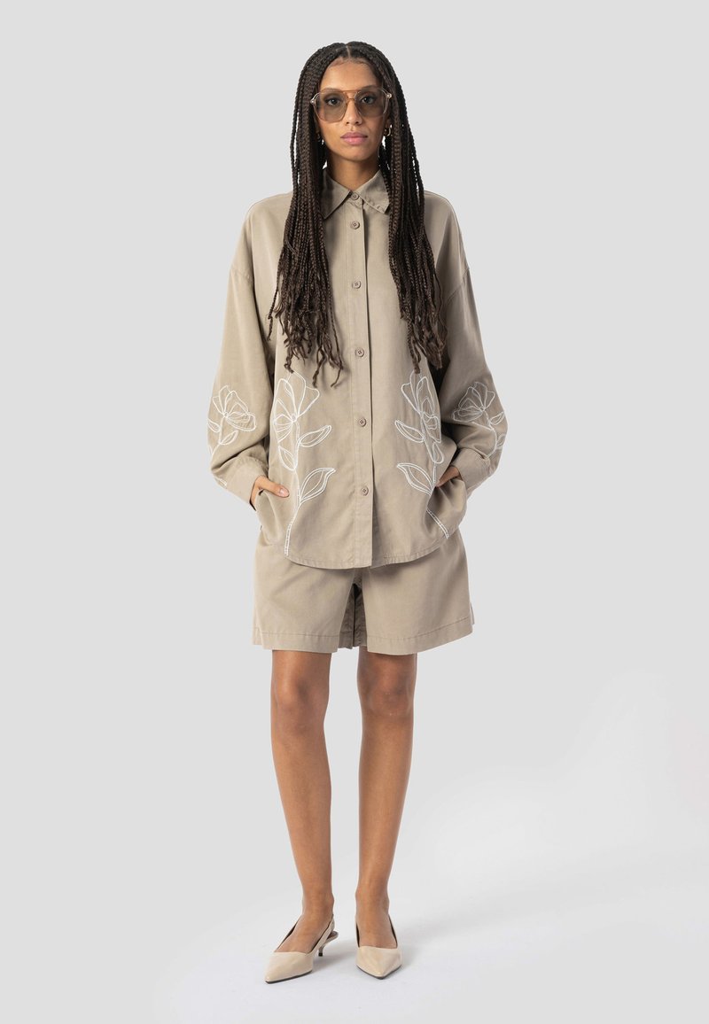 Femme aux longs cheveux tressés portant une chemise beige surdimensionnée avec broderies florales blanches, un short assorti, des talons beiges et des lunettes de soleil.