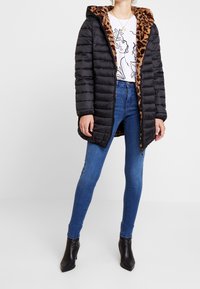 Abrigo tipo puff negro con forro y capucha de leopardo, con cierre de cremallera y diseño acolchado, combinado con vaqueros azules y botas negras de tobillo.