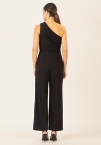 Jumpsuit nero con design a spalla unica, gambe larghe e texture del tessuto morbida. Presenta pieghe in vita e scarpe con tacco alto.