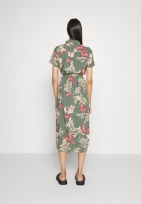 Abito midi floreale in tessuto verde con motivi di farfalle e foglie rosa e beige, maniche corte e vita con cintura. Calzature nere.