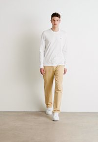 Camisa blanca de manga larga y pantalones caqui, complementados por zapatillas blancas. Diseño simple, con detalles mínimos y un ajuste relajado.