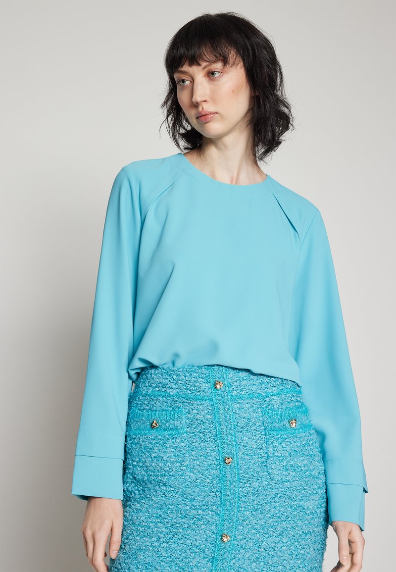 Helle blaue Bluse mit langen Ärmeln und runder Ausschnitt, kombiniert mit einem strukturierten, leuchtend blauen Rock mit goldenen Knopfakzenten.