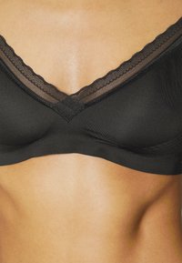 Crni bralette s dizajnom u obliku slova V, s čipkastim obrubom i glatkim materijalom. Mekana tekstura s minimalnim detaljima od tvrdog materijala.