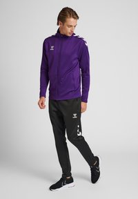 Giacca sportiva viola con zip e tasche laterali, adornata con logo bianco del marchio e strisce sulle spalle. Pantaloni neri con accenti bianchi e logo.