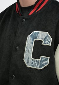 Μαύρο πανωφόρι varsity με γιακά με κόκκινες ρίγες, κουμπιά με κλικ και μεγάλο μπλε πατς με γράμμα «C» με σχέδιο στο στήθος.