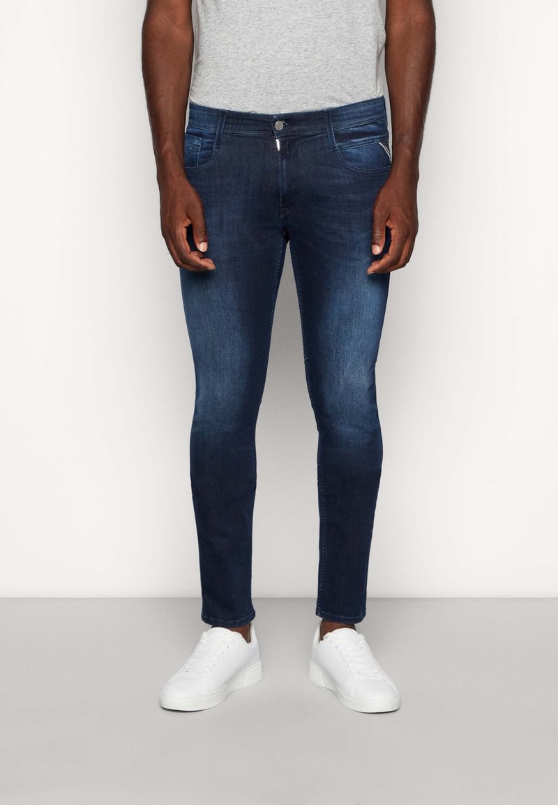 Homme portant un jean bleu foncé ajusté et des baskets blanches, debout devant un fond clair uni.