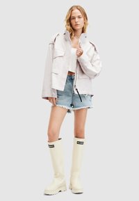 Chaqueta corta de color púrpura claro con dos bolsillos frontales, combinada con pantalones cortos de mezclilla desgastados. Botas blancas hasta la rodilla con marca negra en la parte superior.