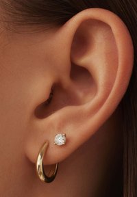 Boucle d'oreille en or et boucle d'oreille à clou avec une pierre claire ronde, présentées sur la peau, mettant en valeur les textures métalliques et réfléchissantes.