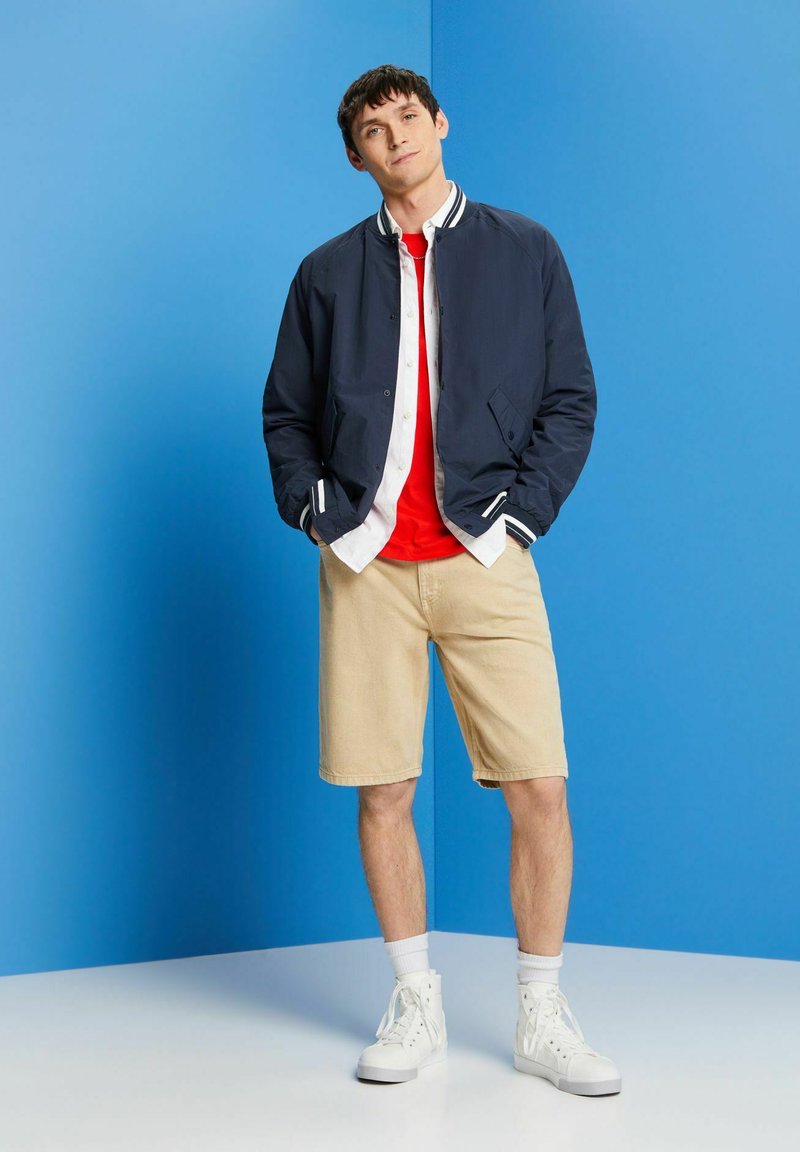 Veste de bombardier marine avec des poignets rayés blancs, T-shirt rouge, shorts crème, et baskets montantes blanches, sur un fond bleu.