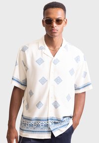 Chemise à manches courtes, boutonnée, en crème avec des motifs géométriques bleus et une bordure florale. Fabriquée en tissu léger, elle offre une coupe décontractée.
