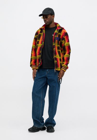 Raudona, geltona ir juoda tikrinto rašto vilnos džemperis su užtrauktuku, derinamas su juodu marškinėliu ir laisvomis mėlynomis džinsais. Užsidėtos juodos bateliai ir kepurė.