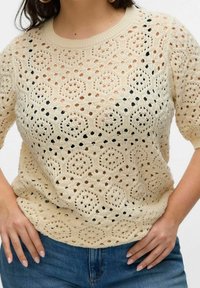 Top corto a maniche corte in crochet beige con motivi circolari, indossato sopra uno strato scuro e abbinato a jeans blu.