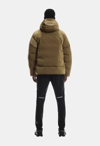 Pufferjacke in olivgrün, mit Kapuze und gesteppter Optik. Getragen mit schwarzen Hosen und Sneakers. Reflexionselemente an den Hosen.