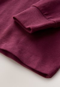 Szczegół burgundowej bawełnianej bluzy z ribbowanym mankietem i dołem. Tekstura wydaje się gładka, a przeszycia podkreślają krawędzie.
