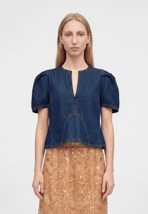 Mujer rubia que lleva una camiseta de manga corta de mezclilla azul oscura con cuello en V y una falda beige con motivos florales con lentejuelas.