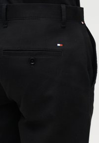 Μαύρο παντελόνι με λεία υφή, με πίσω τσέπη με κουμπί και μια μικρή κεντημένη λεπτομέρεια με σημαία σε κόκκινο, άσπρο και μπλε χρώμα.