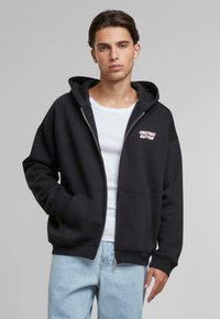 Hanorac negru cu fermoar, din bumbac, cu un buzunar frontal, glugă și logo-ul alb "Another Cotton". Asortat cu blugi de culoare albastru deschis.