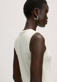 Gebreide mouwloze top in off-white, met een gestructureerde afwerking, ribbeltrim en een hoge hals. Model zijaanzicht.