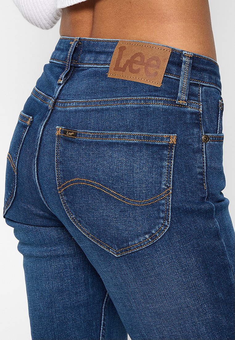 Jean en denim bleu foncé avec une texture lisse, dotés d'un patch en cuir marron affichant la marque "Lee" et de coutures dorées sur les poches.