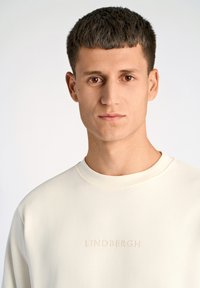Ljus beige sweatshirt med rund halsringning, detaljerad med en präglad "LINDBERGH"-logotyp framtill. Slät textur, avslappnad passform.